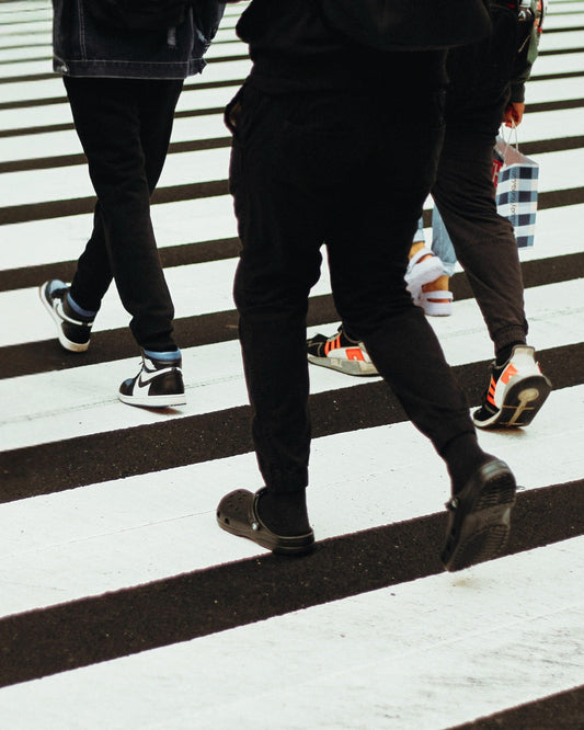 Various people walking across a crosswalk.  Only their feet, legs, and torsos are visible